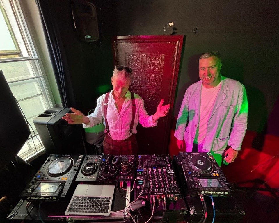 Two DJs behind a full deck setup in the top floor room, lit pink and green